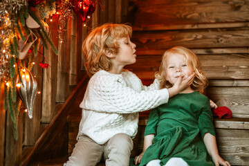 siblings children boy and little girl with blond hair and blue eyes in beautiful clothes sit in living room on stairs near Christmas tree for family celebration of Christmas and New Year, kiss and hug