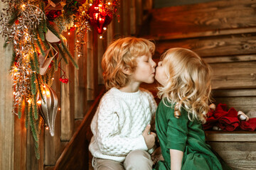 Fototapeta na wymiar siblings children boy and little girl with blond hair and blue eyes in beautiful clothes sit in living room on stairs near Christmas tree for family celebration of Christmas and New Year, kiss and hug