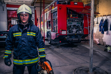 A fireman with uniform and helmet holding a chainsaw with fire truck in the background