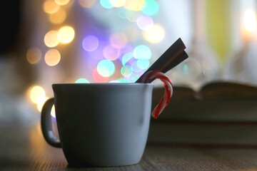 Cup of warm drink with cinnamon stick and candy cane, open book, reading glasses, lit candles and colorful bokeh lights. Selective focus.
