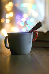 Cup of warm drink with cinnamon stick and candy cane, open book, reading glasses, lit candles and colorful bokeh lights. Selective focus.