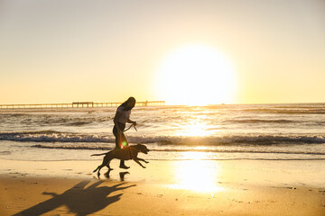 Young Woman Running Behind Dog At Beach