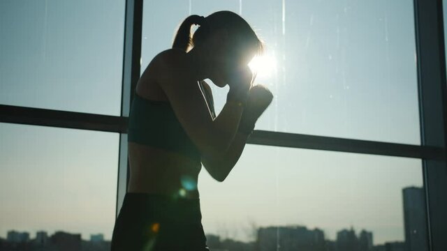 Backlit silhouette of slim young woman throwing punches working out in modern gym against panoramic window. People and sports concept.