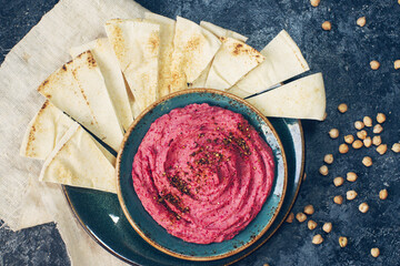 Homemade beetroot hummus, chickpeas and pita slices in blue dish on dark stone table