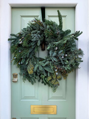 Christmas mood. Elegant christmas wreath decorated fir tree, grass and botanical details on light green wooden door