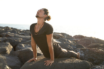 Yoga teacher is practicing on the rocks, sunset time. Man exercising, stretching body. Pranayama. Mindfulness. Meditation.