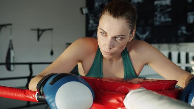 Slow motion portrait of exhausted female boxer resting in ring corner wiping sweat from face with glove looking at camera feeling tired but motivated