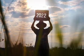 Silhouette of a senior farmer standing in a rice field at sunset and holding a New Year 2022 sign.