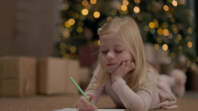 Little Girl Of Four Years With Blond Long Hair. Behind It Christmas Tree Sparkles With Garlands. Girl Draws. She Is On Floor Near Christmas Tree. Near Christmas Tree Gifts. She Has Thoughtful Look.