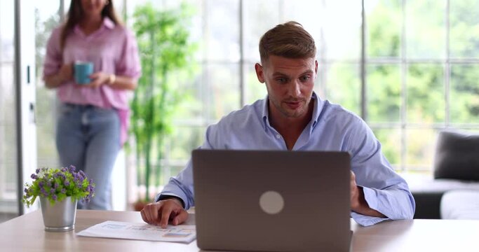 Millennial Caucasian serious busy tired angry moody businessman in casual wear video call teleconference with colleagues on laptop computer while wife bring cup of coffee serving to calm husband down.