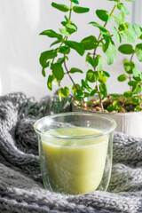 Hot green tea matcha latte on the window wrapped in a warm gray wool scarf of chunky knit. Whipped foam green tea in a glass transparent cup. atmosphere of comfort and winter hygge