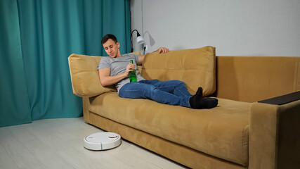 White robot vacuum cleaner delivers alcohol beer bottle to brunet short-haired man sitting on sofa in grey t-shirt and jeans in living room.