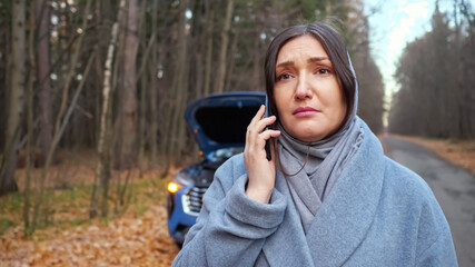 Lady driver in warm coat stands near broken automobile with open hood and calls emergency repair service for help on roadside near autumn forest, closep