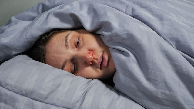 Young brunette woman with runny nose can not fall asleep because of severe cough and lies covered from head to toe with warm blanket closeup.