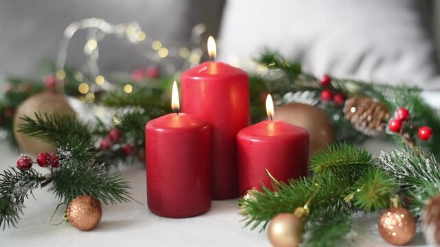 Christmas decoration with three red candles, spruce branches, cones, balls and garland on white table on blurry background. Side view. Hand of woman moves one candle. New year mood, festive concept.