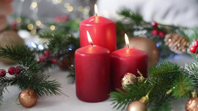 Christmas decoration with red candles, spruce branches, cones, balls and garland on white table on blurry background. Side view. Hand of woman puts one candle. New year mood, festive concept