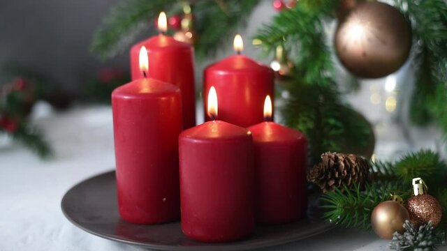 Christmas decoration with red candles on brown plate, spruce branches, cones, balls and garland on white table on blurry background. Side view. New year mood, festive concept, holiday table, gift card