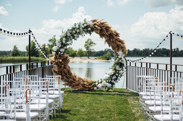 Round arch for a wedding ceremony by the river. White glass chairs stand for guests