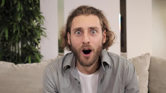 Oh my god wow Amazed excited young man opens his eyes wide sitting on the couch and surprised looking at camera open mouth, shocked by sudden victory. Incredible human reaction to victory at home.