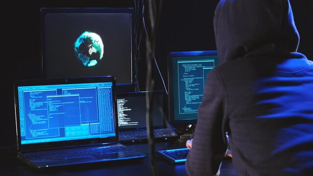 Back hooded hacker using malicious software hack corporate data center. malefactor hidden underground in dark place, multiple displays with phishing code and global map attack.