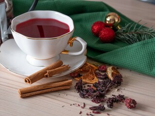 Christmas pink flavored tea in a white cup and saucer. Dry rose petal tea with pieces of fruit and berries is scattered on the table. Green napkin, spruce twig, Christmas balls.