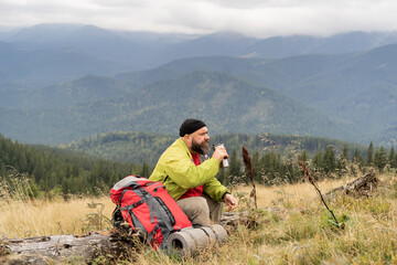 Fototapeta premium a bearded Caucasian male tourist on vacation while hiking in the mountains sits on the trunk of a fallen tree and drinks tea or coffee from a thermos cup. Halt in the mountains.