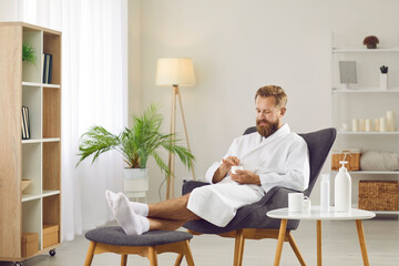Male beauty. Cheerful bearded man enjoys cosmetic procedures at home by applying moisturizing mask on his face. Funny man in bathrobe and white socks sits on chair and tries out various cosmetics. - obrazy, fototapety, plakaty