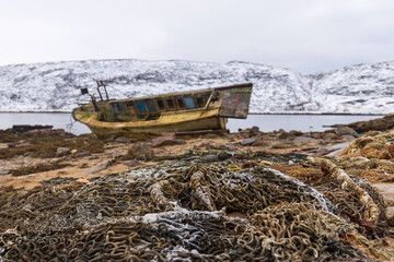 Fototapeta na wymiar A pile of old fishing nets covered with snow on the sand against the background of an old broken fishing boat. Teriberka village, Murmansk region, Russia.