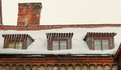 The roof of an old red brick church with a chimney and three windows covered with snow and icicles.
