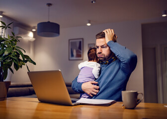 Remote work difficulties and fatherhood. A nervous young father holding his sleepy baby girl and having troubles with his job. A man holds his head and looks at the laptop in disbelief.