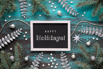Winter background, text Happy Holidays. Xmas border with fir twigs, Christmas decoration baubles and exotic fern leaves and light garland. Acrylic background in turquoise, brown. Flat lay, top view.