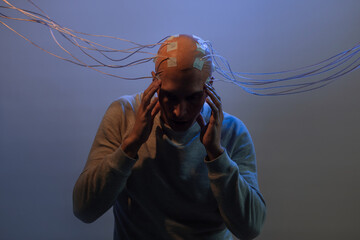 A young man with his head entangled in wires, clutched his head, electrical contacts on his bald head, signals to the brain. Neurointerface, future technologies, creative concept.
