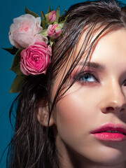 Fototapeta na wymiar A young beautiful woman with rose flowers in her wet hair. Portrait of a beautiful brunette with perfect skin, close-up on a blue background