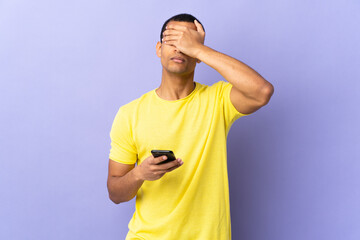 African American man over isolated purple background using mobile phone covering eyes by hands