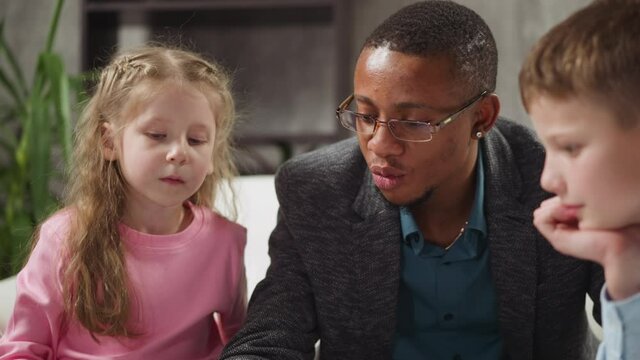 Black man English native speaker writes explaining lesson topic material to attentive little siblings at private tuition at home slow motion