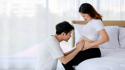 Asian young happy loving and supporting husband sitting and lovely look to unborn baby in beautiful pregnancy wife naked big belly tummy stomach sitting on bed in bedroom at home