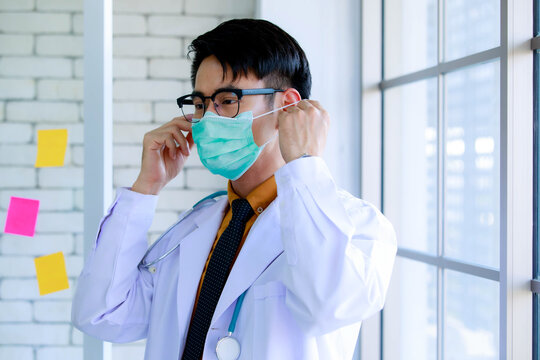 Close Up Shot Of Asian Successful Professional Male Doctor Wear Eyeglasses In White Lab Coat Uniform Hanging Stethoscope Around Neck Standing Wearing Safety Hygiene Facial Mask In Hospital Office