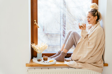 Happy blond woman warming and cover knitted plaid enjoying in her coffee time by the window in cold winter day. Peace of mind and mental health.