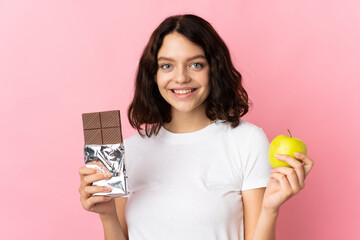 Teenager Ukrainian girl isolated on pink background taking a chocolate tablet in one hand and an apple in the other