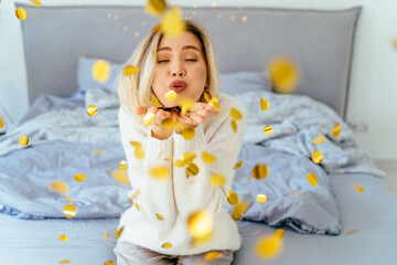 Caucasian blond e woman blowing confetti while sitting on bed at home.