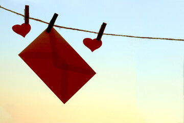 Love letter envelope and two red hearts hang on rope attached with clothes peg pins over orange morning sky sunrise background. Moment of romance love, Happy Valentine Day celebration.