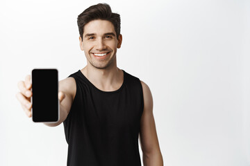 Handsome strong guy showing smartphone screen and smiling. Male athlete demonstrating workout app...