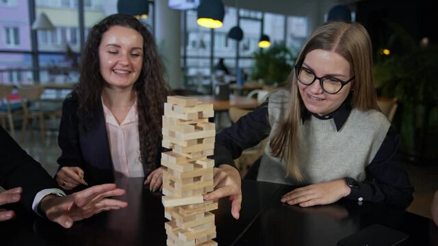 Company of friends playing jenga and having fun. One of the girls carefully pushes the brick in the pile of jenga. Leisure time for adults.