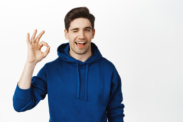 Fototapeta premium Handsome young man winks and shows okay sign, confirms smth, recommends place, stands over white background