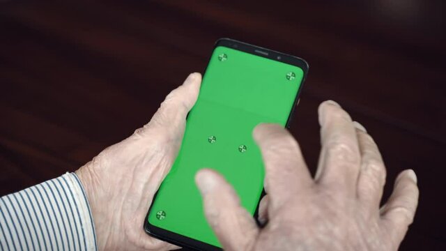 Close up shot of senior man hands holding trendy smartphone with green screen tracking mockup and scrolling pages with finger, lonely man in retirement spending his free time browsing internet pages