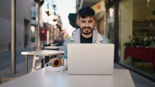 Young hispanic man listening to music sitting on table at coffee shop terrace
