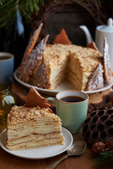 Napoleon cake decorated with Christmas gingerbread cookies. Fir branches, cones, candles, coffee, side view, wooden background.