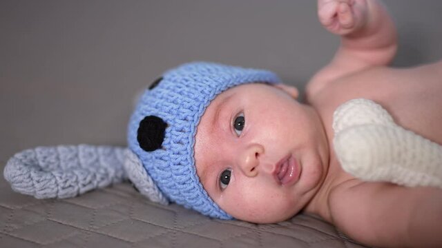 Small kid lying and moving his tiny hands quickly. Adorable babe in funny blue doggy cap and knitted bone. Beautiful child in the bedroom.