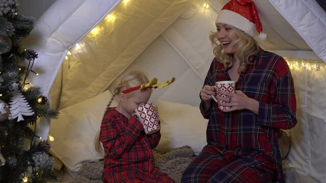 Mom and child drink cocoa at home in the Christmas interior. People in pajamas on New Year's Eve