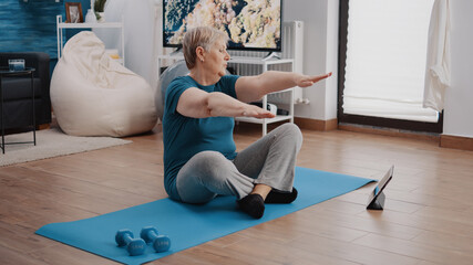 Elder woman following training lesson program on tablet while doing physical exercise and workout. Retired adult looking at gadget screen to practice aerobics and fitness activity. - obrazy, fototapety, plakaty